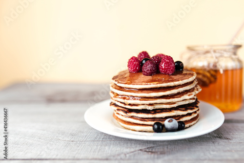 Juicy pancakes with berries and honey on a white plate, spoon, jar, wooden table