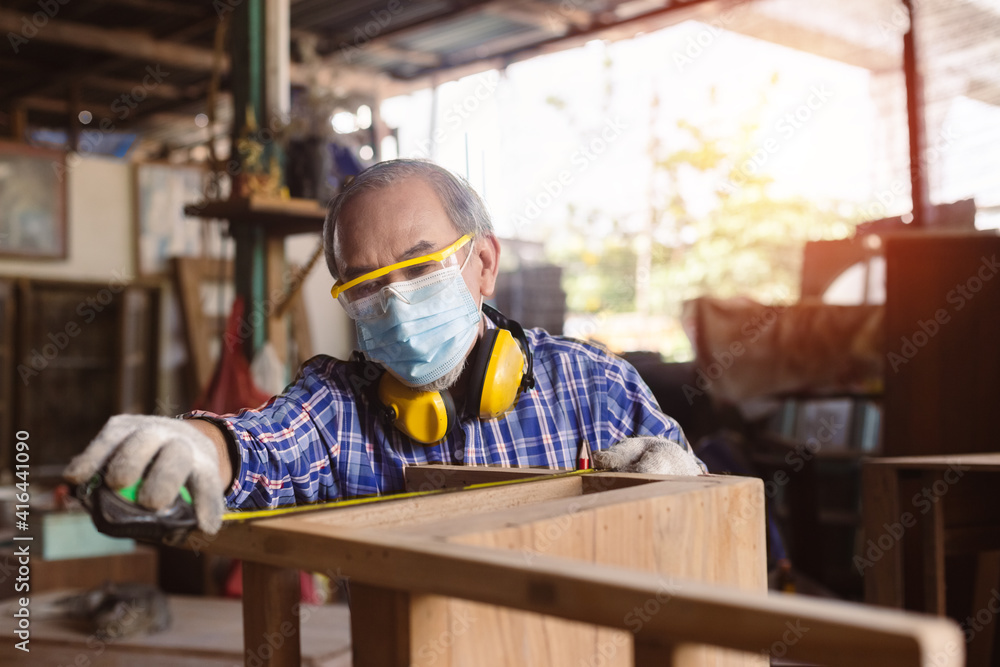 asian senior man carpenter wear protective mask dust and headphone working use measuring tape looking wood size at workspace. craftsman profession in wood factory.