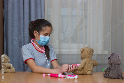 A girl with dark school-age hair, wearing a gray T-shirt, medical mask, plays in the hospital with toys, vaccinations.
