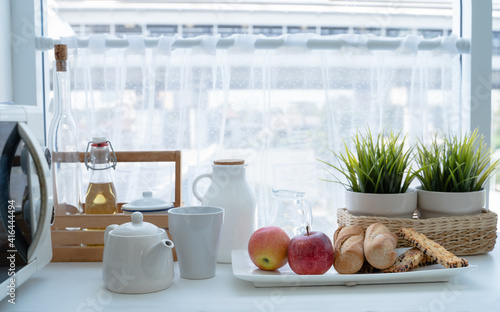 Breakfast in the white kitchen. red apples, loaves of bread, pastry, tea pot, bottle of milk and honey on the table next to window in the morning