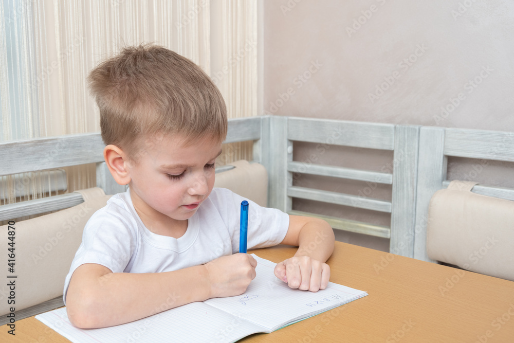 The child studies online at home. Cute little boy writes homework in a notebook sitting at the table at home. Online home education concept The child studies online at home. Cute little boy writes homework in a notebook sitting at the table at home. Online home education concept