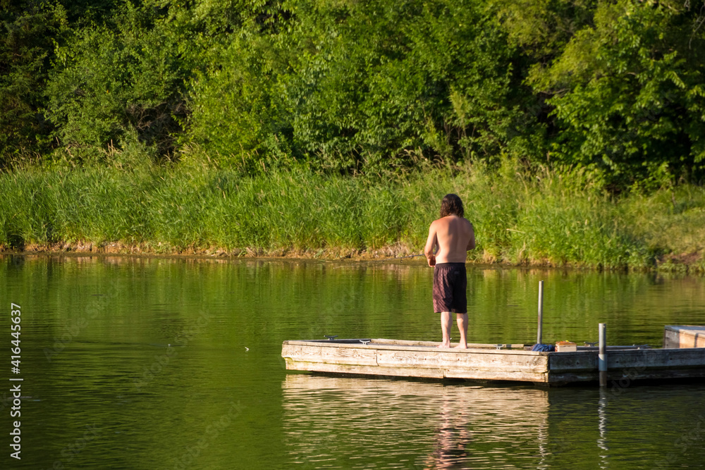A shirtless man fishing off a wooden deck on a lake on a hot summer day.