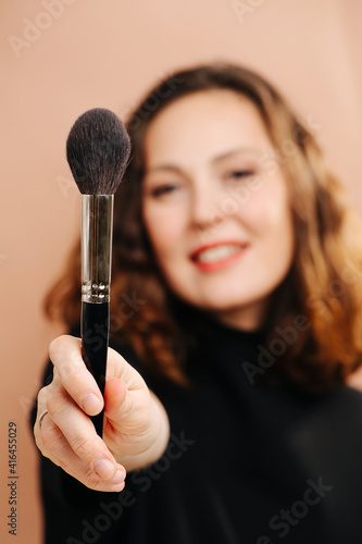 Smiling woman with blurred face showing her cosmetic brush, holding it in front of the camera. Selective focus on the object.