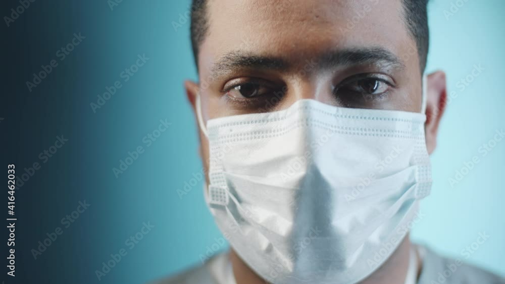 Close up studio portrait of middle eastern male doctor in medical face mask posing for camera on blue background during covid-19 pandemic Close up studio portrait of middle eastern male doctor in medical face mask posing for camera on blue background during covid-19 pandemic