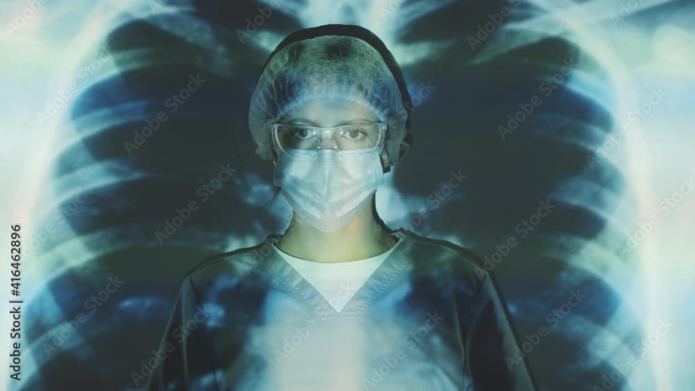 Portrait shot of young female doctor in medical uniform, protective glasses, mask and disposable hat standing in front of projection screen with chest x-ray and looking at camera Portrait shot of young female doctor in medical uniform, protective glasses, mask and disposable hat standing in front of projection screen with chest x-ray and looking at camera