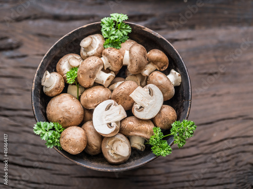 Brown colored common mushrooms in wooden bowl on wooden table with herbs. Top view.