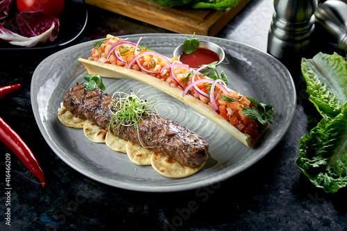 Appetizing Turkish lamb lula kebab with tomato tartare and red sauce, served in a gray plate. Dark marble background. Barbecue meat, restaurant food.