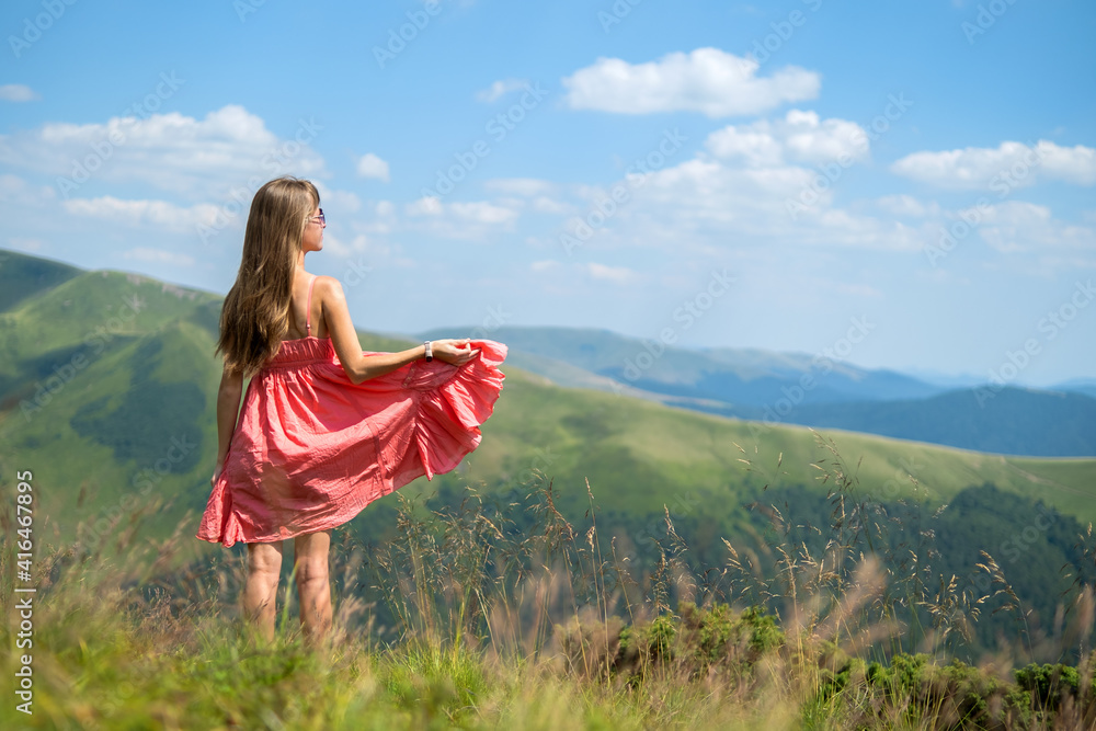 Young woman in red dress standing on grassy field on a windy day in summer mountains enjoying view of nature.