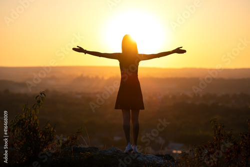A young woman in summer dress standing outdoors with outstretched arms enjoying view of bright yellow sunset.