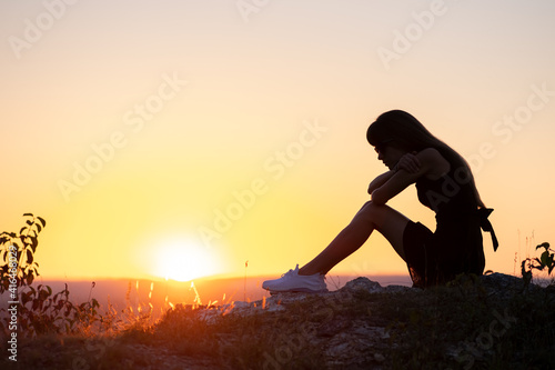 Young depressed woman in black short summer dress sitting on a rock thinking outdoors at sunset. Fashionable female contemplating in warm evening in nature.