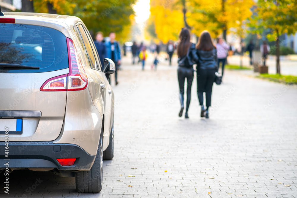 Cars parked in a row on a city street side on bright autumn day with blurred people walking on pedestrian zone. Cars parked in a row on a city street side on bright autumn day with blurred people walking on pedestrian zone.