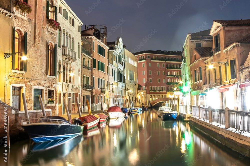 Fototapeta premium Evening view of illuminated old architecture, floating boats and light reflections in canals water in Venice, Italy.