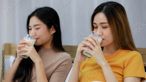 Smiling Two Asian young womenDrink milk in a glass lovely couple on white bed and happy. Funny women Before going to bed together on cozy. Concept In love homosexual lesbian lgbt, lgbtq, lgbtq+.