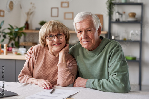 Portrait of senior couple embracing and smiling at camera while sitting at the table with laptop and documents at home