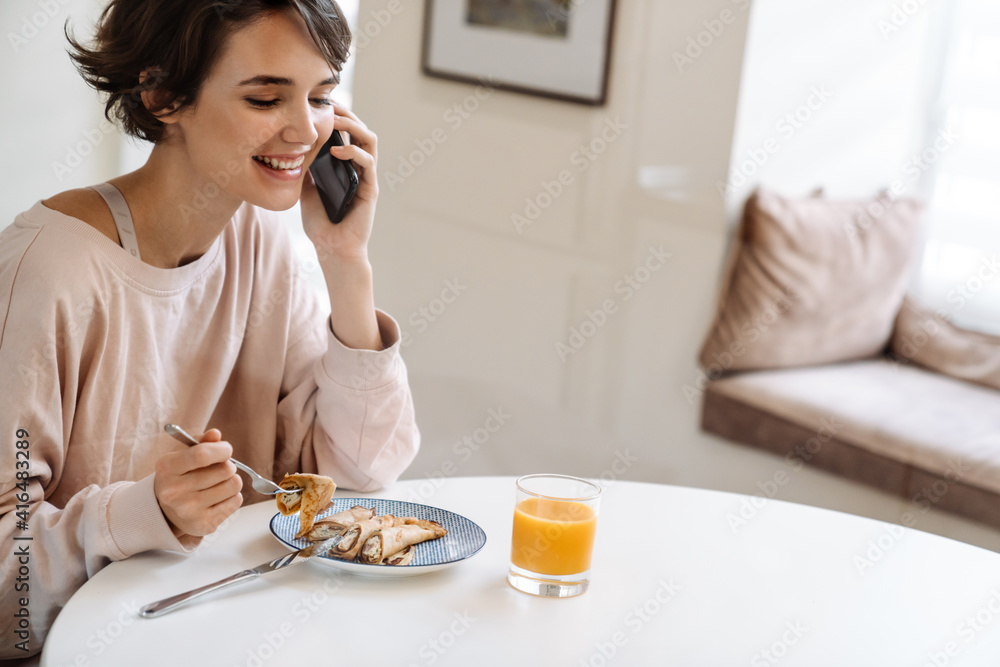Joyful beautiful girl talking on cellphone while having breakfast Joyful beautiful girl talking on cellphone while having breakfast