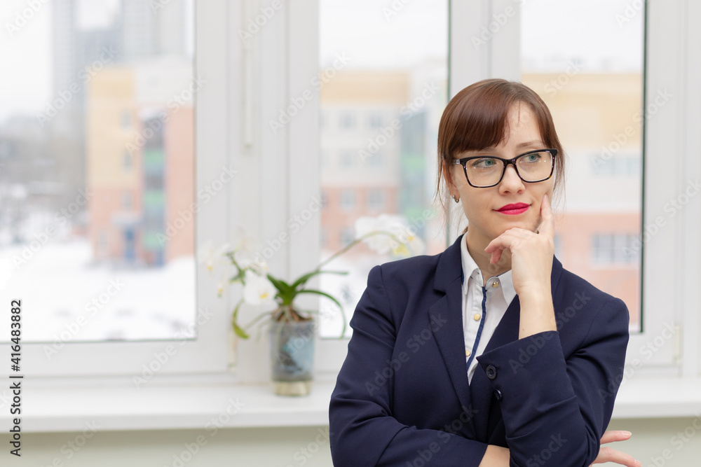 young stylish pretty business woman in a blue jacket and glasses by the window in the office at her workplace is satisfied with the work done young stylish pretty business woman in a blue jacket and glasses by the window in the office at her workplace is satisfied with the work done