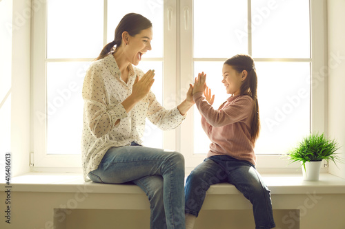 Happy family having fun at home. Mom and little child spending time together. Young mother and cute daughter playing patty cake sitting on windowsill of plastic window back lit with bright sunlight photo