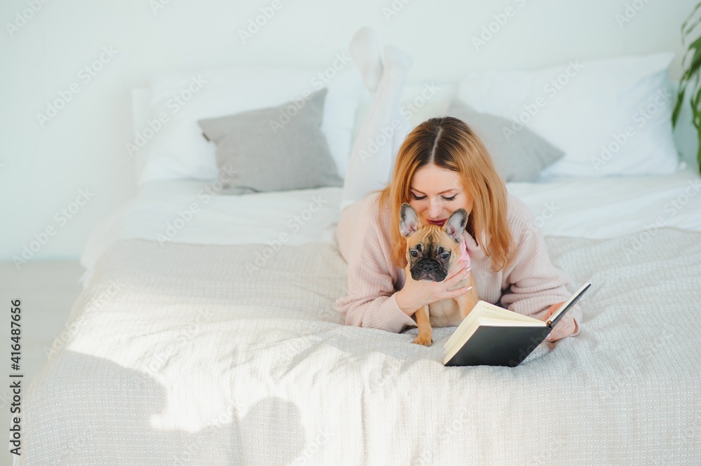 A woman reading a book and smiling as she sits in bed. The alarm clock on the desk beside her. A woman reading a book and smiling as she sits in bed. The alarm clock on the desk beside her.