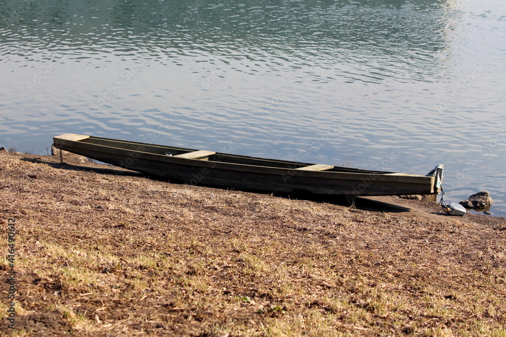 Dilapidated old wooden river boat made from wooden boards left on dry grass covered river bank next to calm river on warm sunny winter day