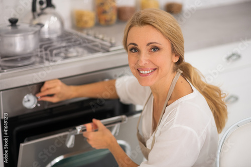 Blonde woman opening the oven in the kitchen while cooking dinner