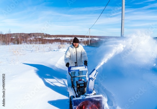 A man in a white jacket removes snow from a rural road with a blue snowblower in winter after a snowfall