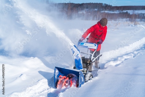 A woman in a red jacket removes snow from a rural road with a snowblower in winter after a snowfall photo