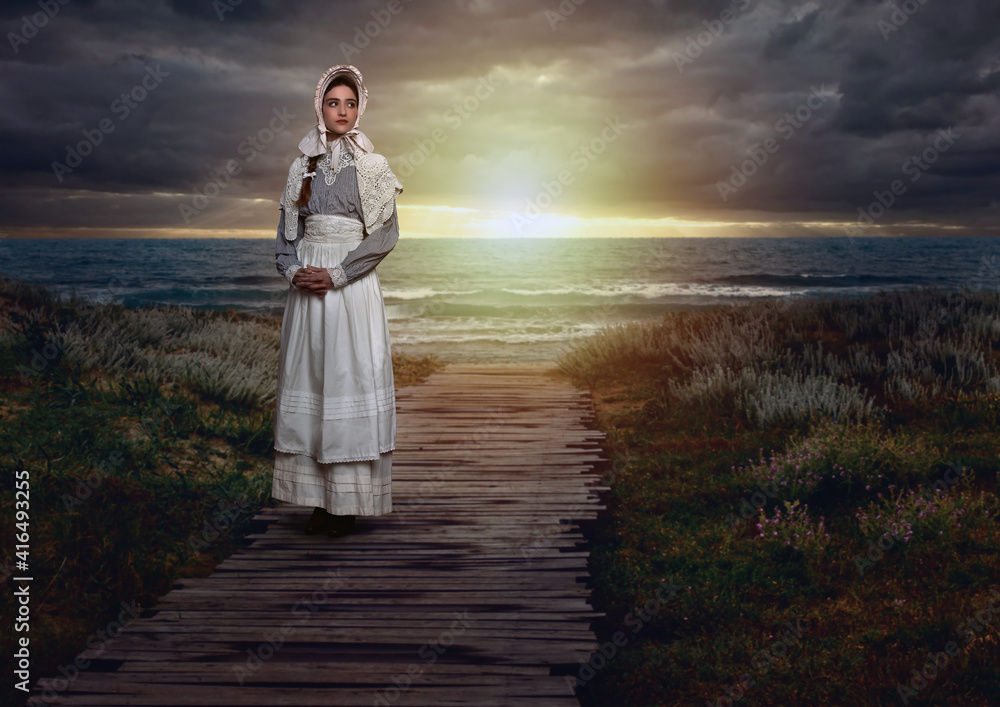 Victorian girl with a cap on in a white dress and blue striped blouse on the coast at sunset.