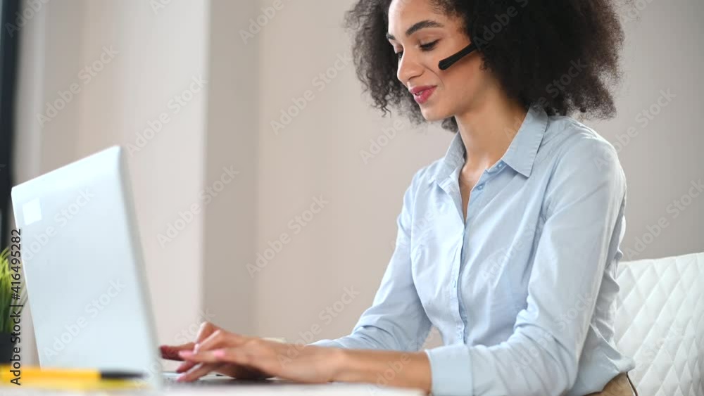 A focused mixed-race businesswoman with Afro hairstyle in headset sitting at the desk in the modern office and working on a laptop, video-calling, providing information and help to clients A focused mixed-race businesswoman with Afro hairstyle in headset sitting at the desk in the modern office and working on a laptop, video-calling, providing information and help to clients