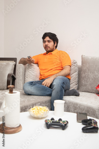 Asian Indian man gaming, streaming and watching TV alone in living room on a grey sofa and white table