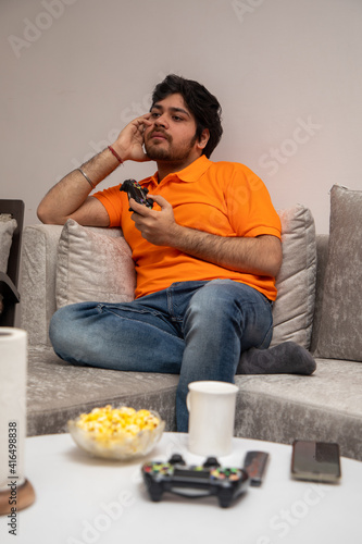 Asian Indian man gaming, streaming and watching TV alone in living room on a grey sofa and white table