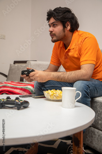 Asian Indian man gaming, streaming and watching TV alone in living room on a grey sofa and white table