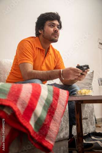 Asian Indian man gaming, streaming and watching TV alone in living room on a grey sofa and white table