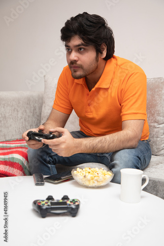 Asian Indian man gaming, streaming and watching TV alone in living room on a grey sofa and white table
