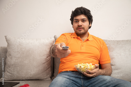 Asian Indian man gaming, streaming and watching TV alone in living room on a grey sofa and white table