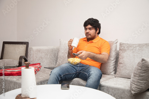Asian Indian man gaming, streaming and watching TV alone in living room on a grey sofa and white table