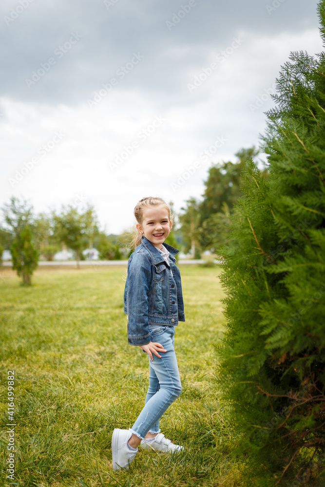 Little smiling girl in jeans jacket posing outdoors in spring park at green bush background. Happy optimistic child, carefree childhood concept