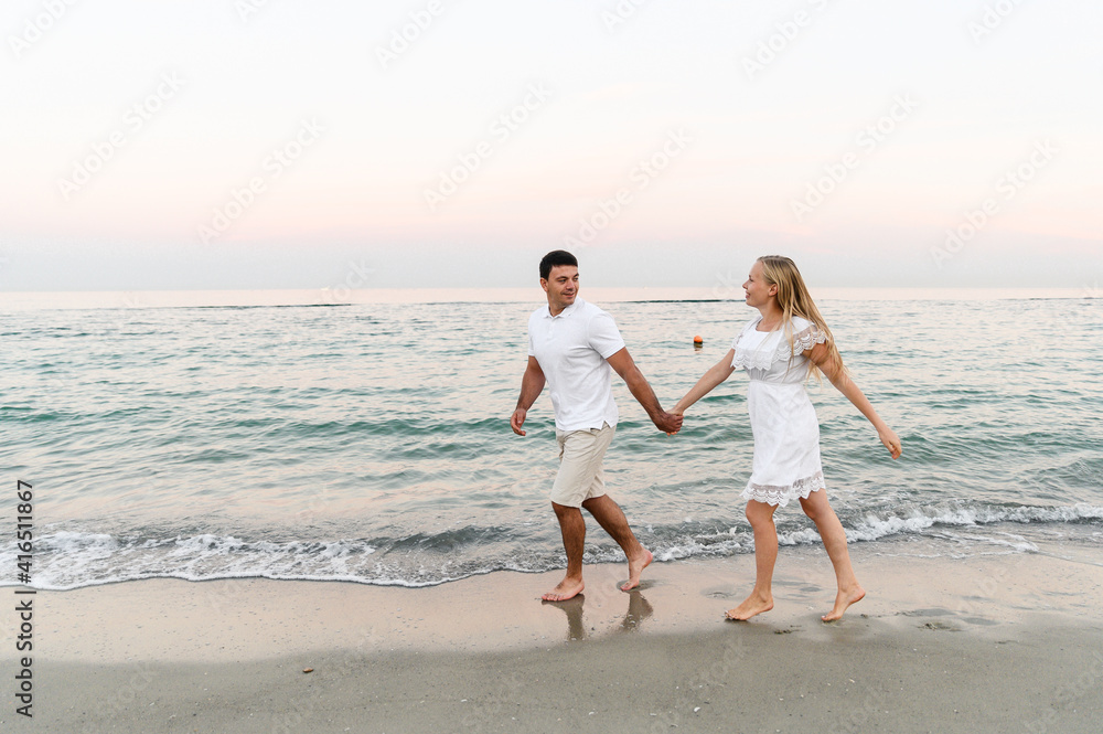 a guy with a girl in summer clothes are walking by the ocean. husband and wife at a romantic sunset of the day near the sea. lovers run along the beach. honeymoon for the newlyweds.