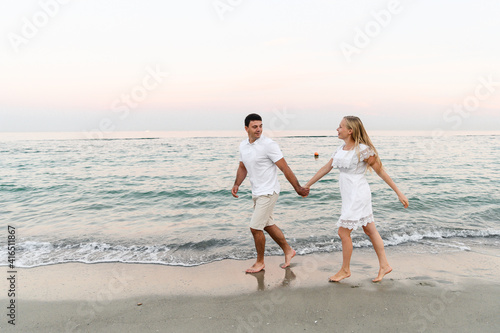 a guy with a girl in summer clothes are walking by the ocean. husband and wife at a romantic sunset of the day near the sea. lovers run along the beach. honeymoon for the newlyweds.