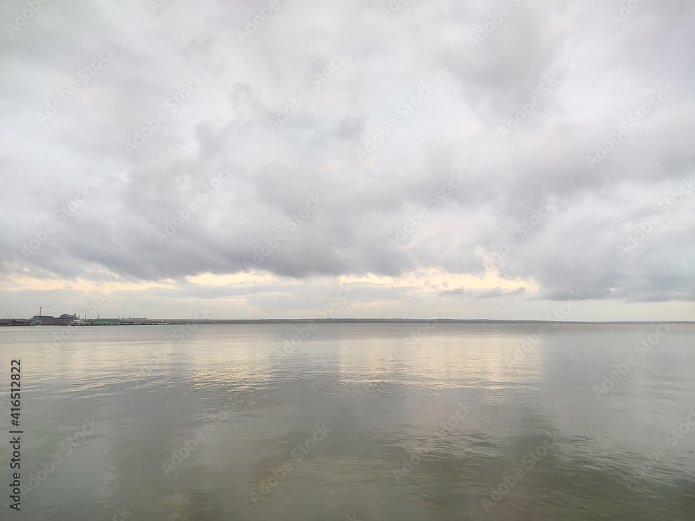 The water surface of the reservoir on a rainy cloudy day. Landscape in dark gray tones. Cumulus clouds hang over the bay. Sunlight and rays barely break through. Horizontal photography.