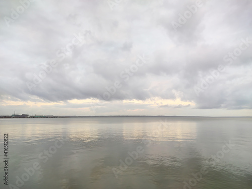 The water surface of the reservoir on a rainy cloudy day. Landscape in dark gray tones. Cumulus clouds hang over the bay. Sunlight and rays barely break through. Horizontal photography. © Oksana Zhigulina	