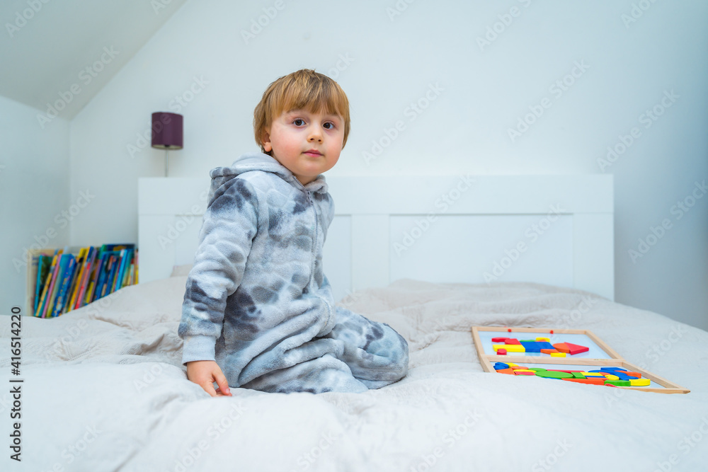Little toddler boy plays on the bed in a wooden magnetic constructor, preschool child learning at home to make mosaic. Early development. Playing with chikldren during coronavirus outbreak. Little toddler boy plays on the bed in a wooden magnetic constructor, preschool child learning at home to make mosaic. Early development. Playing with chikldren during coronavirus outbreak.