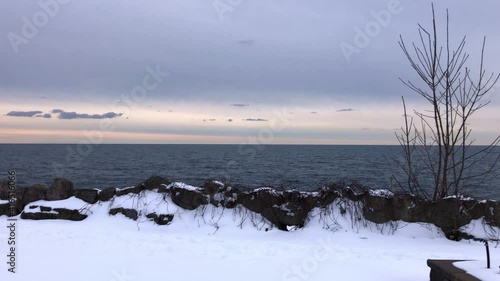 Behind a snow covered chess table, a calm blue lake and pale sky is revealed on a winter day. | 4k, 24fps photo