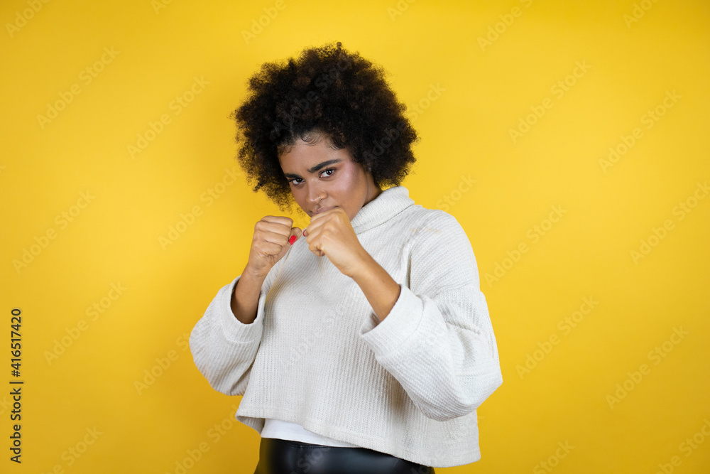 African american woman wearing casual sweater over yellow background Punching fist to fight, aggressive and angry attack, threat and violence African american woman wearing casual sweater over yellow background Punching fist to fight, aggressive and angry attack, threat and violence