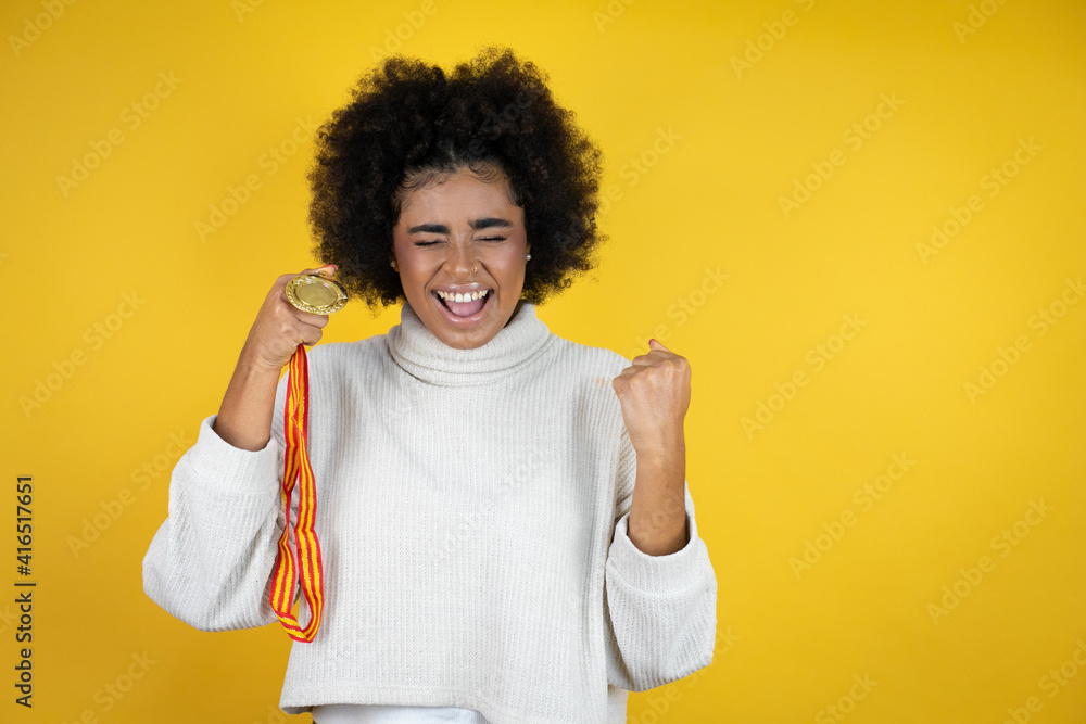 African american woman wearing casual sweater over yellow background holding a medal very happy and excited making winner gesture with raised arms, smiling and screaming for success.