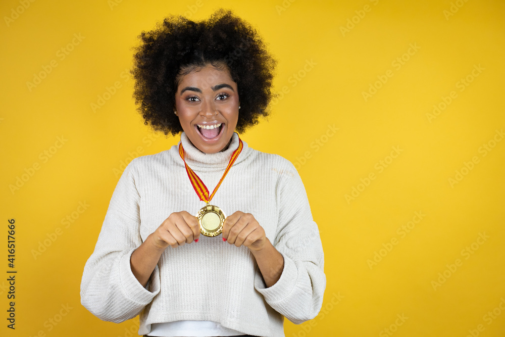African american woman wearing casual sweater over yellow background holding a medal with a happy face standing and smiling with a confident smile showing teeth - obrazy, fototapety, plakaty 