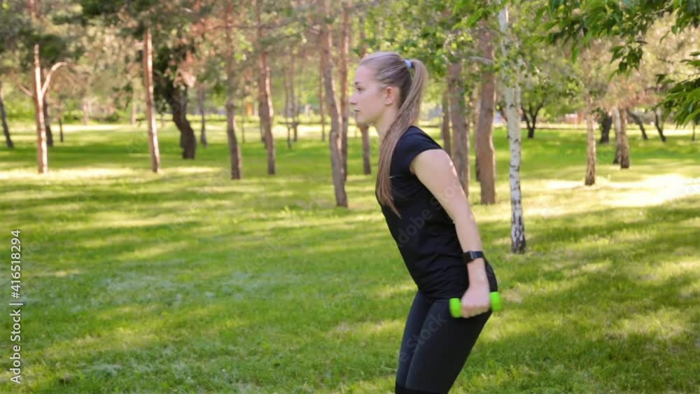An attractive, slim, athletic girl performs exercises with dumbbells for warm-up and arm workout. Fitness in a summer park. An attractive, slim, athletic girl performs exercises with dumbbells for warm-up and arm workout. Fitness in a summer park.