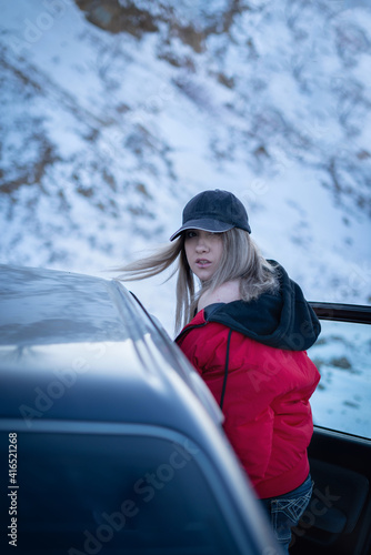 A beautiful girl in a red jacket and black baseball cap with her hair flying in the wind leaned against the car and bared her shoulder sexually. In winter, against the background of snow mountains