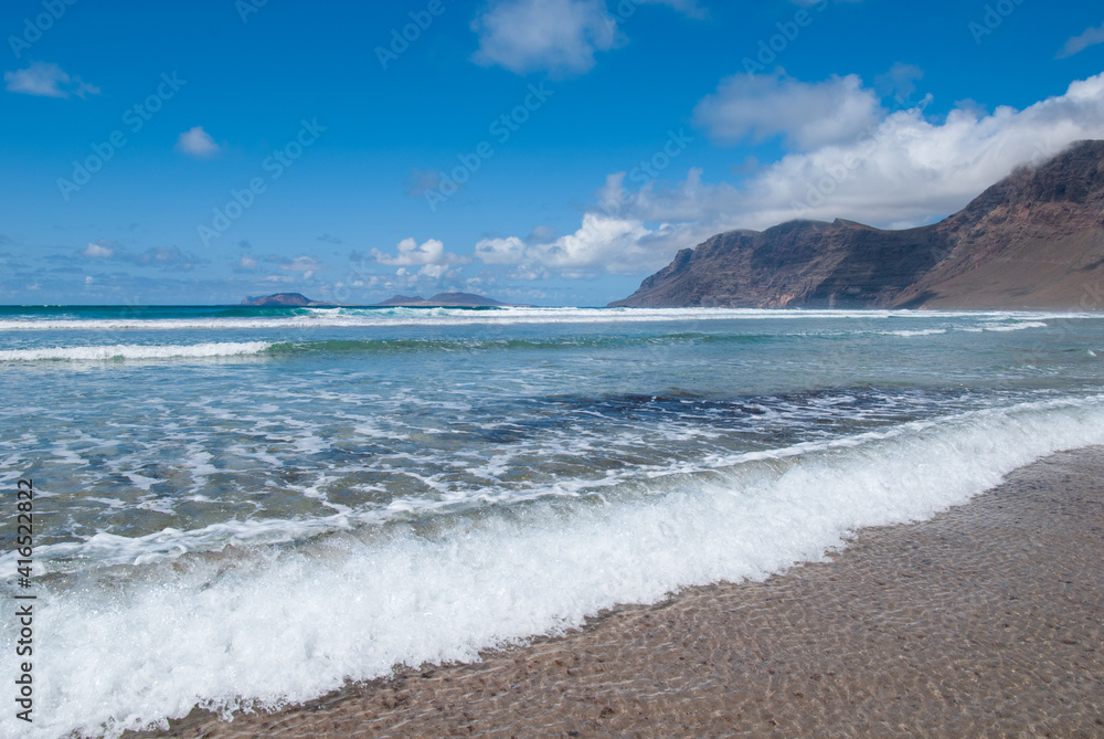 glasklares Wasser spült in Wellen an den Sandstrand Playa de Famara mit Blick auf die Insel La Graciosa, Lanzarote, Kanaren