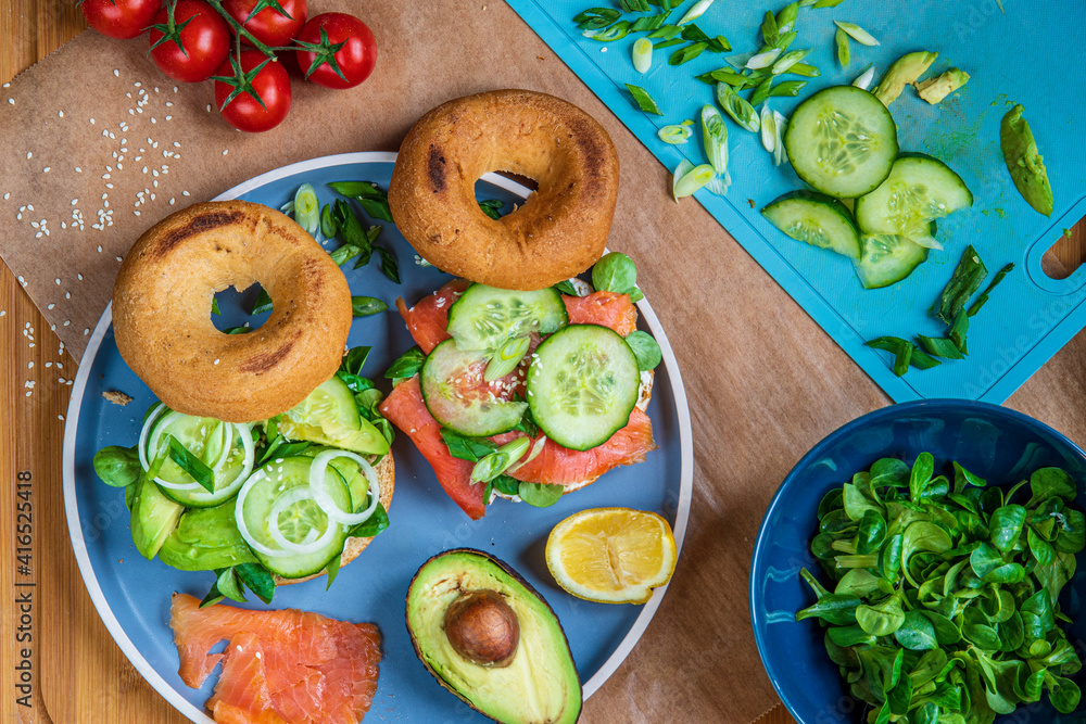 Homemade bagel sandwiches. Gluten free toasted bagels with smoked salmon and cream cheese and with avocado, cucumber, lettuce, and onions. Healthy breakfast on wooden board, fun vibrant colors Homemade bagel sandwiches. Gluten free toasted bagels with smoked salmon and cream cheese and with avocado, cucumber, lettuce, and onions. Healthy breakfast on wooden board, fun vibrant colors
