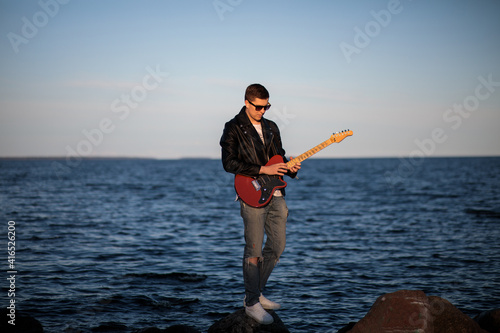 young guitarist in black leather jacket and jeans plays electric guitar against the backdrop of the lake 